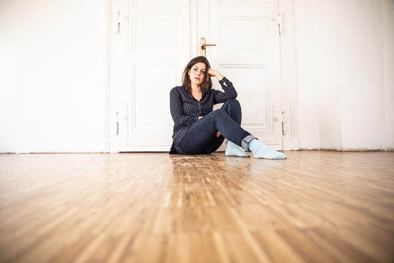 Das Bild zeigt eine Frau in einem fast leerem Raum. Sie sitzt auf dem Boden an die Tür gelehnt und wirkt nachdenklich.
