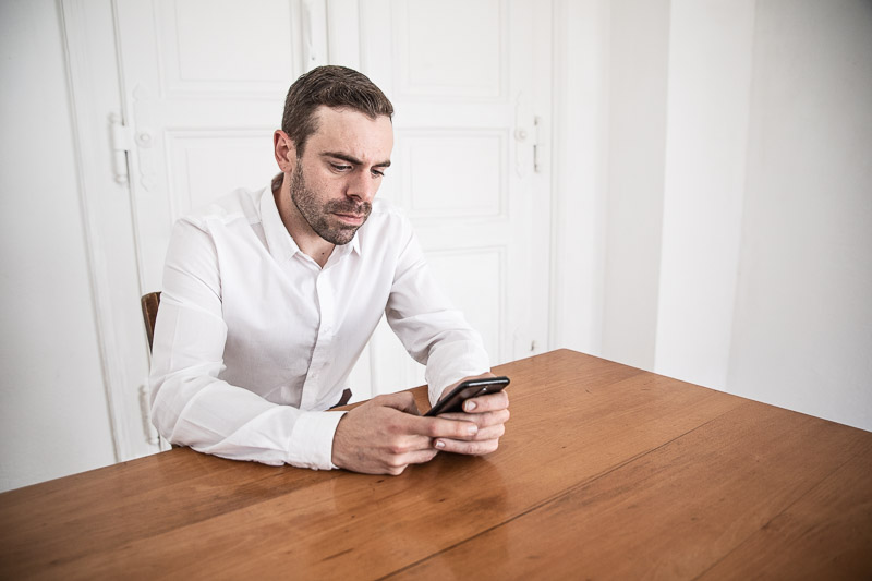 Das Bild zeigt einen Mann an einem Holztisch sitzend. Er schaut auf sein Smartphone.